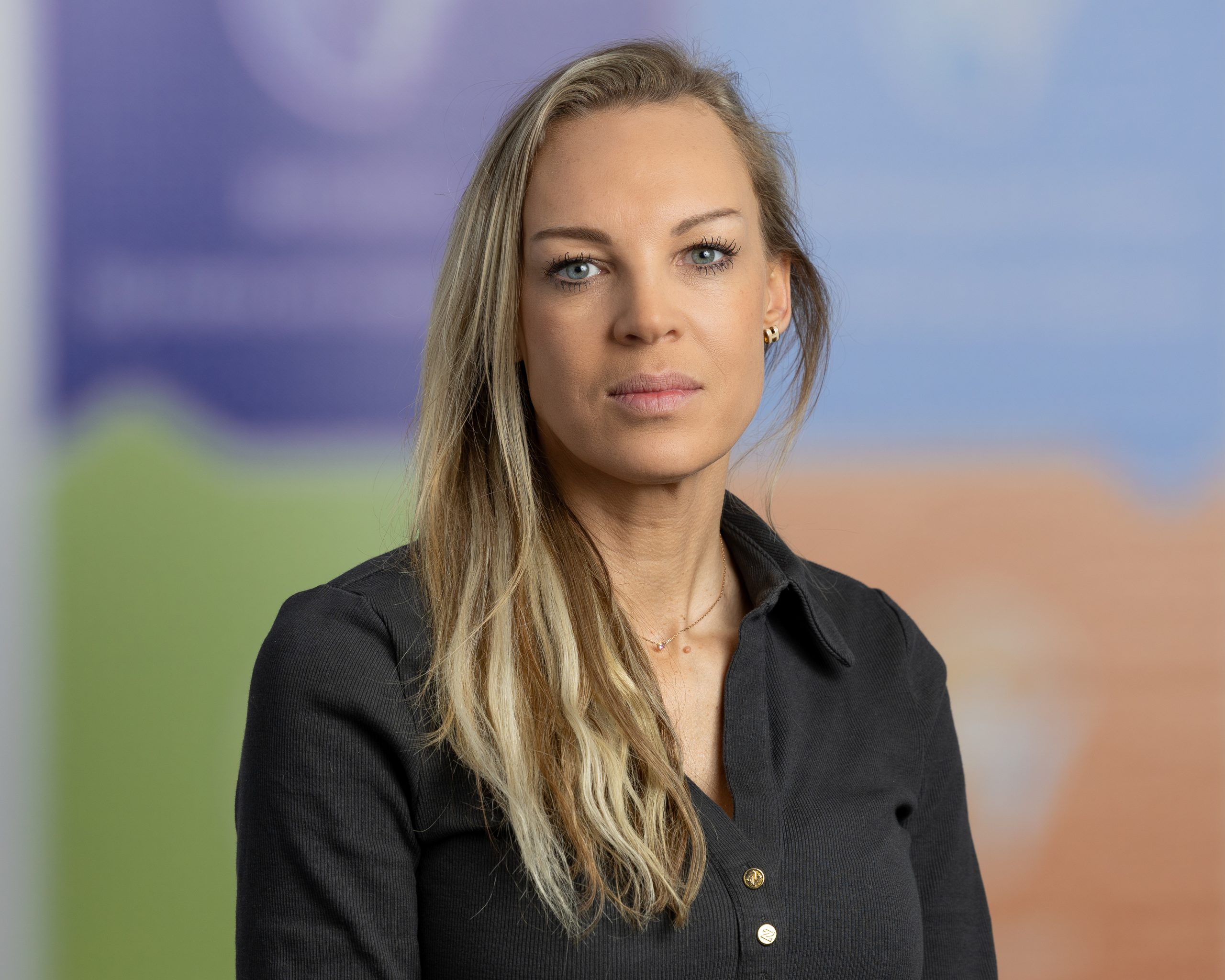 Een vrouw met lang blond haar en een zwart overhemd met knopen kijkt naar de camera tegen een wazige, kleurrijke achtergrond - perfect voor vve beheer of mvgm-professionals die op zoek zijn naar een modern, toegankelijk beeld.